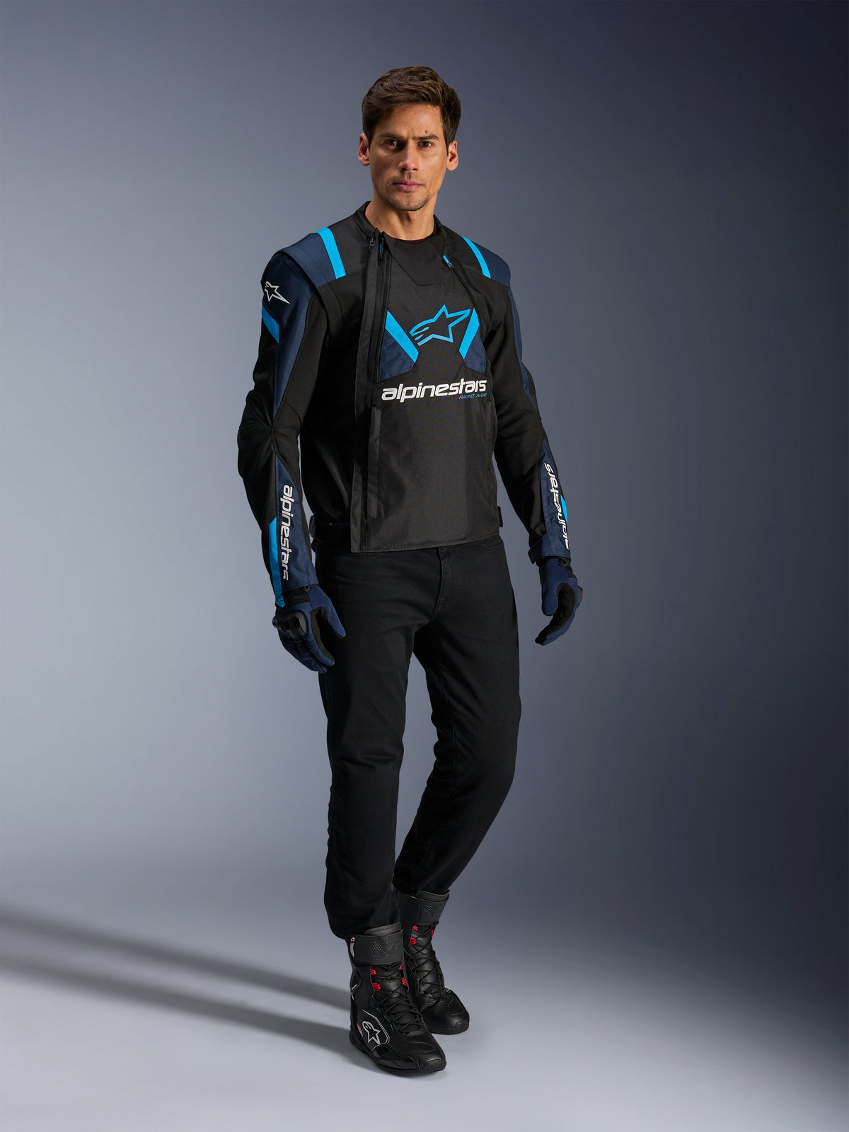 Male model wearing Alpinestars T-Stunt Air Jacket, Black Dark Blue and Blithe Blue, featuring prominent mesh panels for airflow, paired with black pants and riding gloves, sport-cut motorcycle jacket for summer road riding.