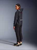Female rider wearing the Alpinestars Molly Air Jacket in Black Fluo Coral, rear side view showing the hood with Astars logo and large Alpinestars branding on the back, paired with black cargo riding pants and tan and black adventure boots, contemporary urban motorcycle gear for women