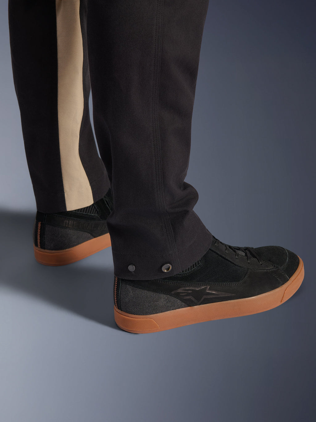 Alpinestars Flex-AST Canvas Pants in Black Aluminum, close-up of lower leg showing snap adjusters at the opening and a vertical aluminum-colored stripe, paired with black riding sneakers with gum soles, urban motorcycle apparel.