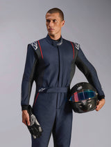 Male model wearing Alpinestars GP Tech V4 Suit, Navy Blue auto racing suit with black and red accents, holding a carbon fiber helmet and racing gloves, professional motorsport protective gear