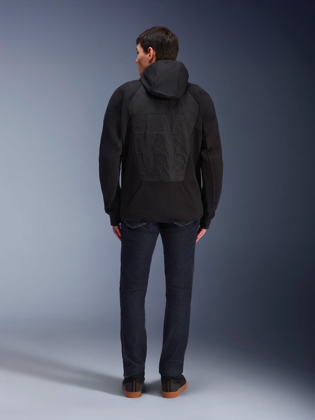 Alpinestars MO.ST.EQ Hybrid Hooded Jacket, Black, rear view showing seamless padded back panel with tonal embossed Alpinestars logo, worn with dark jeans and black sneakers, urban motorcycle streetwear.