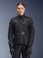Woman wearing Alpinestars Stella ST-7 2L GORE-TEX Jacket, Black with Dark Gray accents, motorcycle touring jacket with large cargo pockets and high collar, paired with black gloves and pants, waterproof adventure riding apparel
