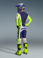 Motocross rider viewed from the back wearing complete racing gear, 2026 Racer Air Riway Pants in Light Gray, Yellow Fluo and Blue, matching jersey with navy blue back and neon yellow accents, coordinated helmet and neon yellow boots, professional motocross outfit.