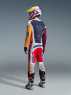 Motocross rider viewed from behind wearing complete racing gear, Alpinestars 2026 Racer Portl Pants in Red Light Gray and Dark Gray, matching jersey with orange and maroon sleeves, multi-colored graphic helmet, gray and black off-road boots with pink highlights, professional motocross outfit