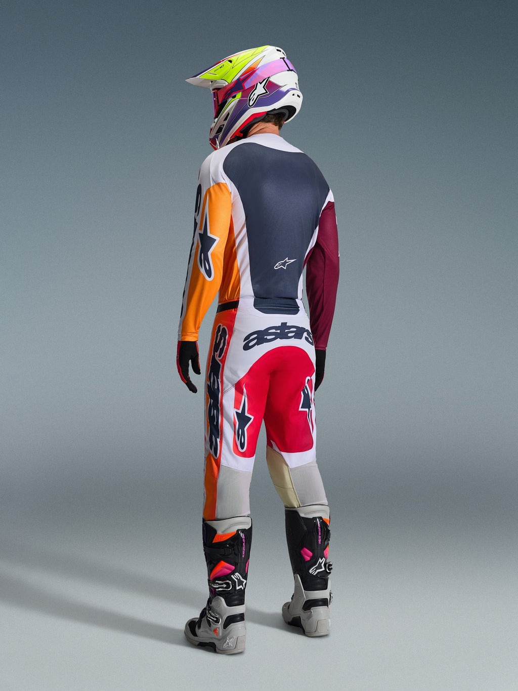 Motocross rider viewed from behind wearing complete racing gear, Alpinestars 2026 Racer Portl Pants in Red Light Gray and Dark Gray, matching jersey with orange and maroon sleeves, multi-colored graphic helmet, gray and black off-road boots with pink highlights, professional motocross outfit