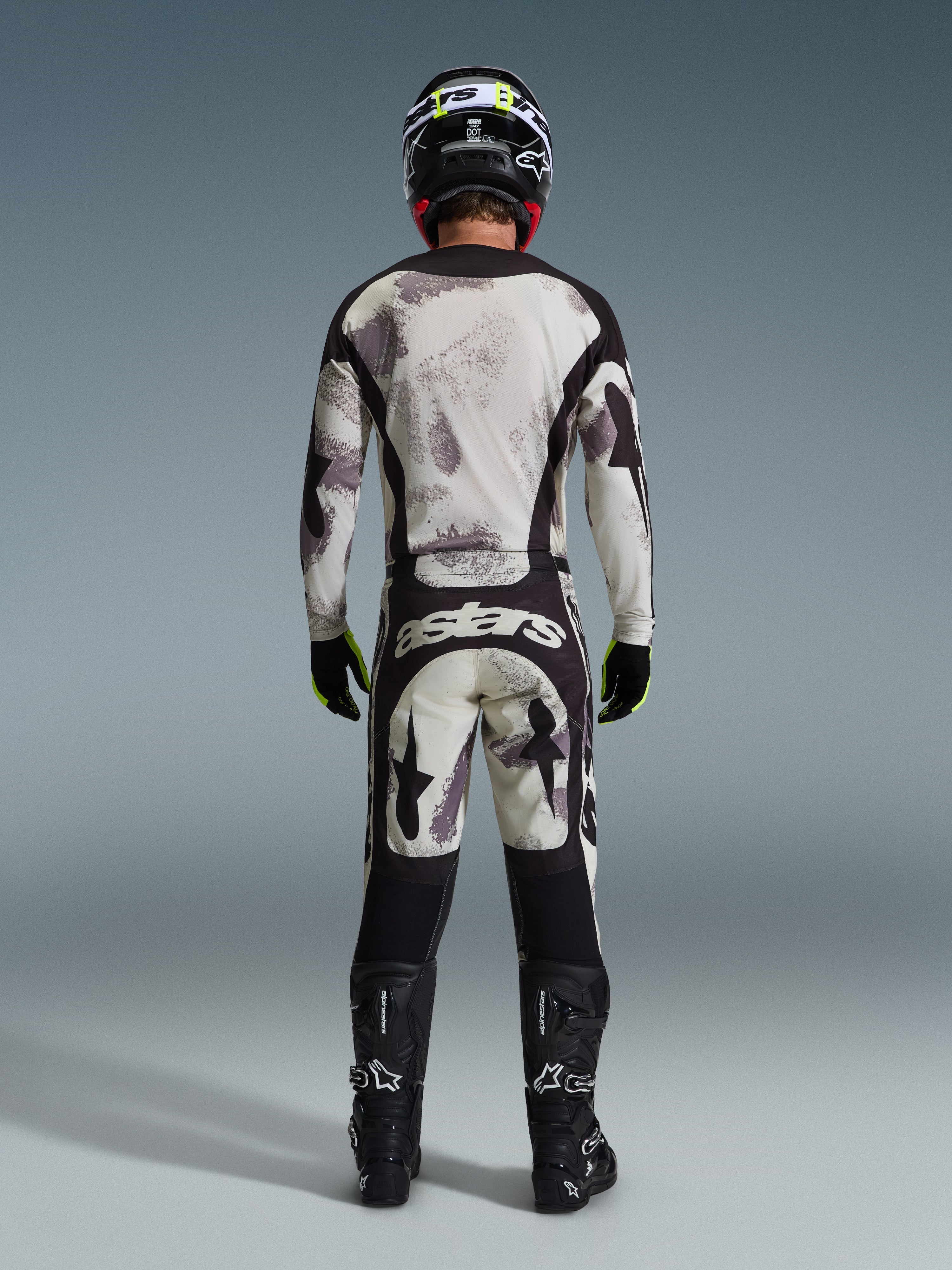 Motocross rider from behind wearing the Alpinestars 2026 Racer Lahnd Pants in Sand Camo, matching long-sleeve jersey with gray camouflage pattern, black motocross boots, black helmet, and "astars" branding on the seat, designed for professional off-road racing.
