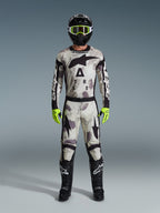 Motocross rider wearing 2026 Racer Lahnd Jersey and matching pants in Sand Camo, black helmet with neon yellow goggles, neon yellow gloves, and black boots, complete Alpinestars off-road racing gear set