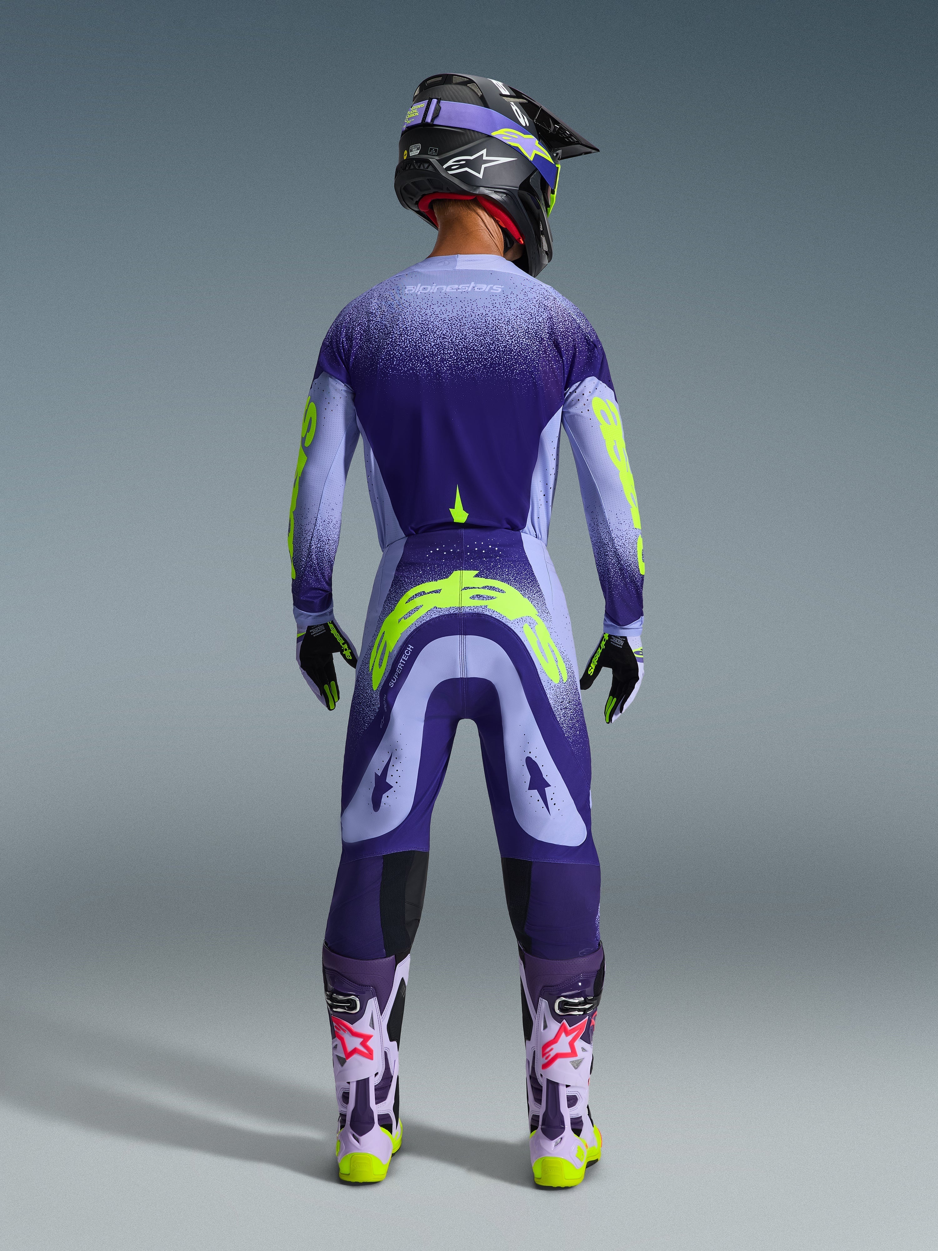 Motocross rider from behind wearing Alpinestars 2026 Supertech Scenz Pants in Purple Yellow Fluo, matching jersey with gradient purple and neon yellow graphics, black helmet, and purple boots, coordinated professional racing gear for off-road performance