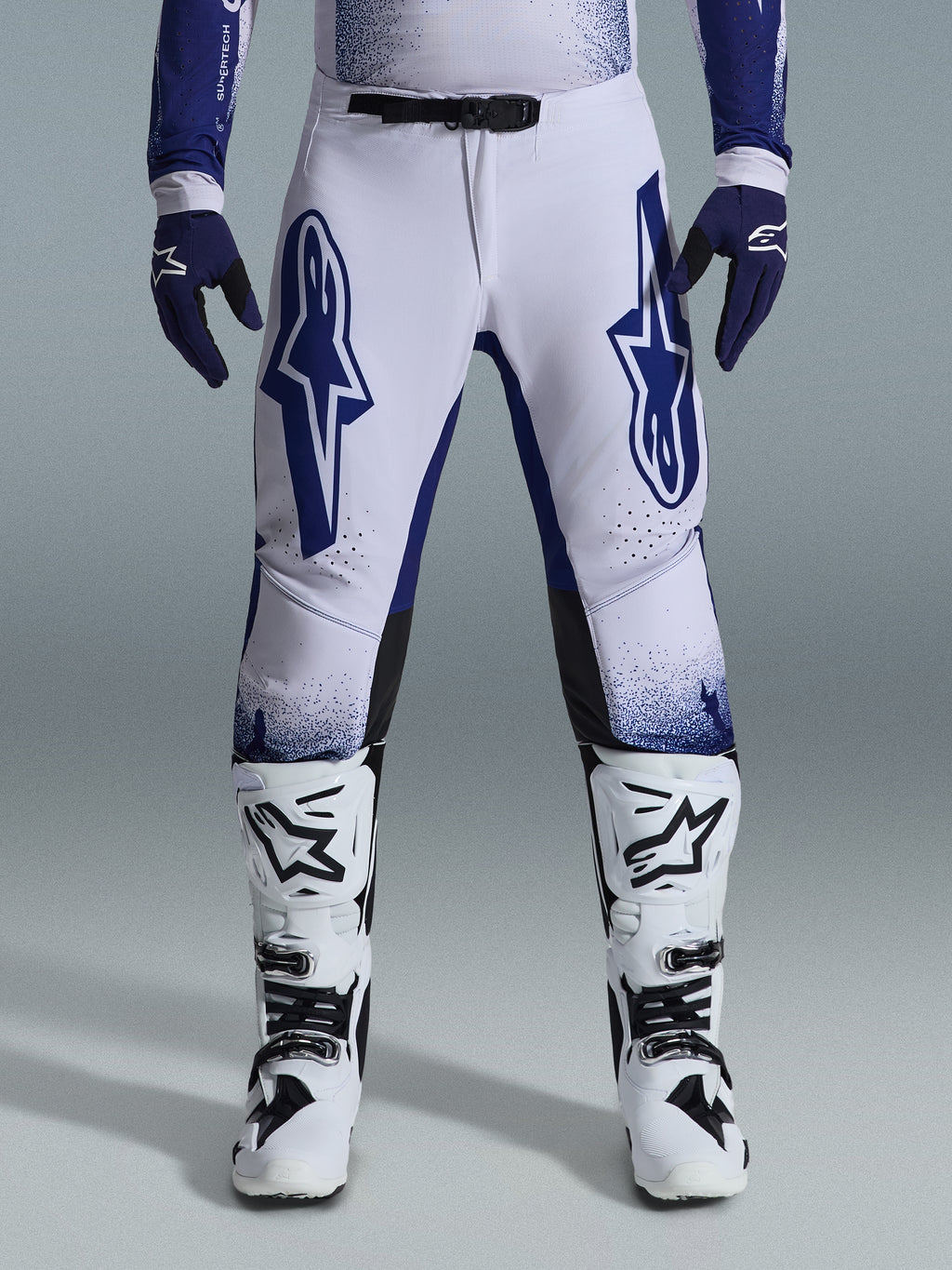 Alpinestars 2026 Supertech Scenz Pants, Motocross Pants, Light Gray and Dark Blue, worn with white and black motocross boots and dark blue gloves, featuring large blue Alpinestars logos on the thighs, SX1 magnetic buckle, and athletic fit for off-road racing