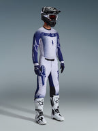 Motocross rider wearing 2026 Supertech Scenz Pants in Light Gray and Dark Blue, matching blue and white jersey with 'astars' branding and spray-paint effect, black helmet with white goggles, white and black motocross boots, coordinated professional off-road racing outfit.