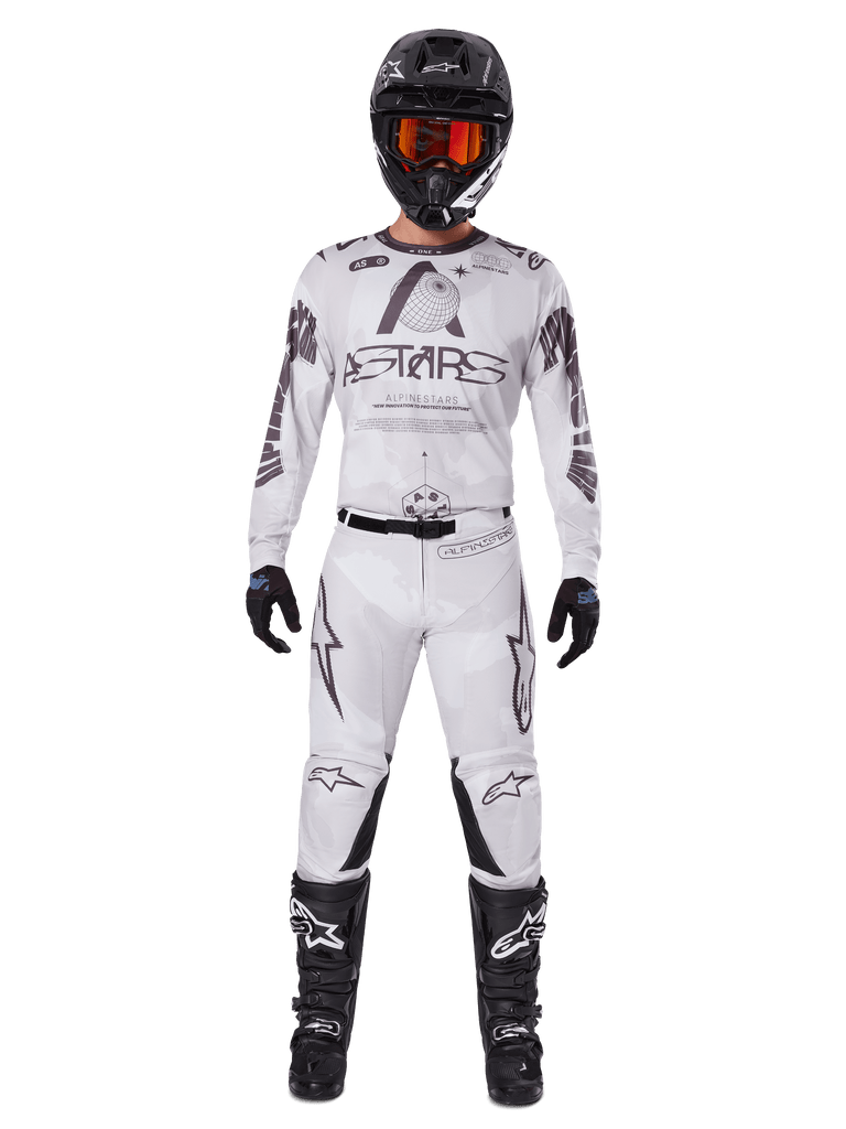 Motocross rider wearing the Alpinestars 2025 Racer Hollow Pants in Light Camo Brown, paired with a matching white and grey long-sleeve jersey, black helmet with orange-red goggles, black gloves, and black boots, coordinated racing gear set for off-road performance.