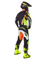 Motocross rider from a rear view wearing the Alpinestars 2025 Fluid Wurx Gear Set, black jersey with yellow and orange accents, matching black pants with white and orange panels, bright yellow boots, and a black helmet, complete off-road racing outfit