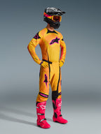 Motocross rider wearing Alpinestars 2026 Techstar Nomur Pants in Orange Yellow Pink, complete racing gear set with matching jersey and gloves, bright pink boots, black helmet with orange goggles, full body view against a grey background, professional off-road racing outfit