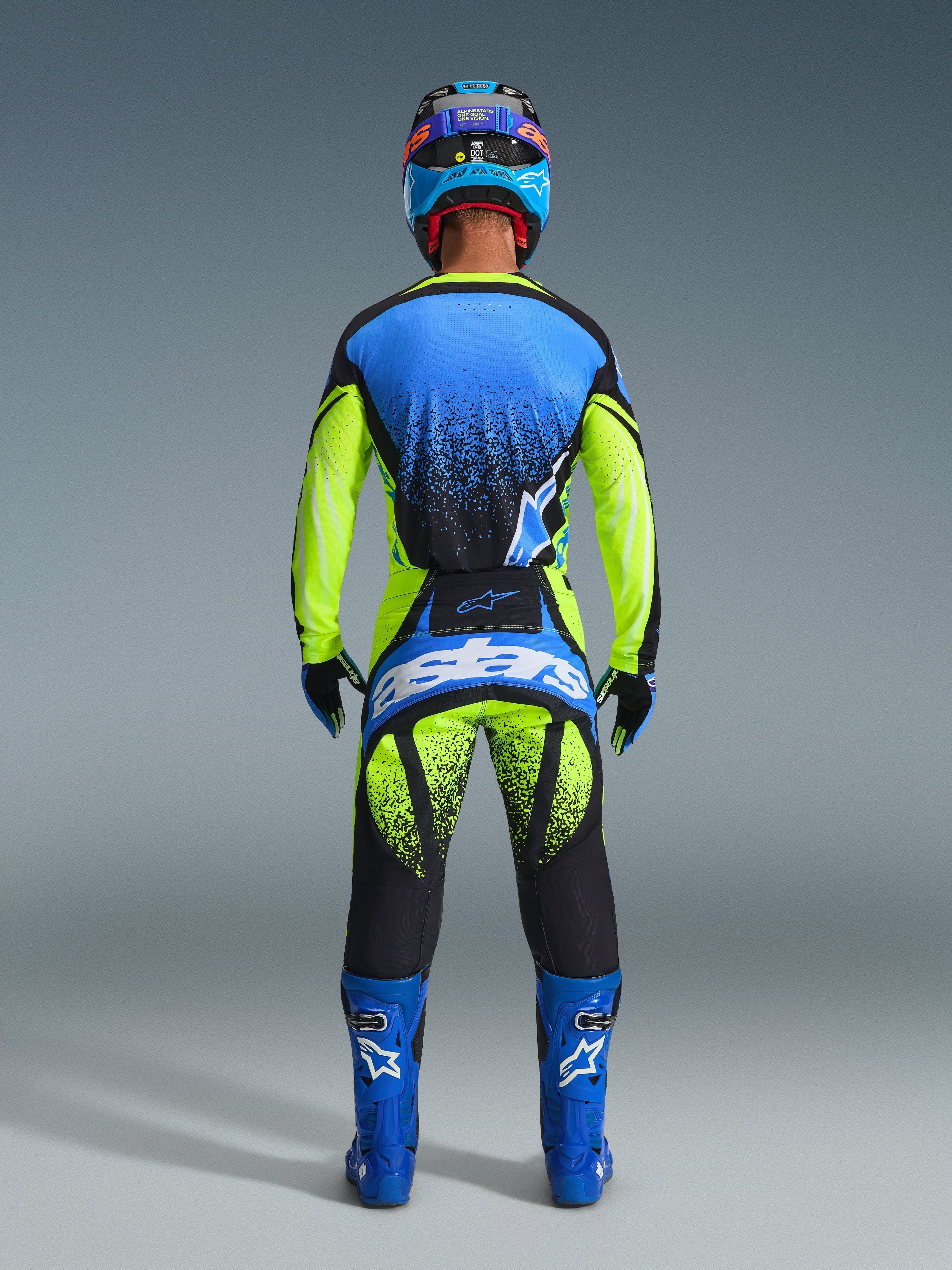 Motocross rider from the back wearing Alpinestars 2026 Techstar Nomur Pants, Yellow Fluo and UCLA Blue with Dark Navy accents, matching jersey and blue boots, featuring bold Astars branding on the seat and a speckled graphic design.