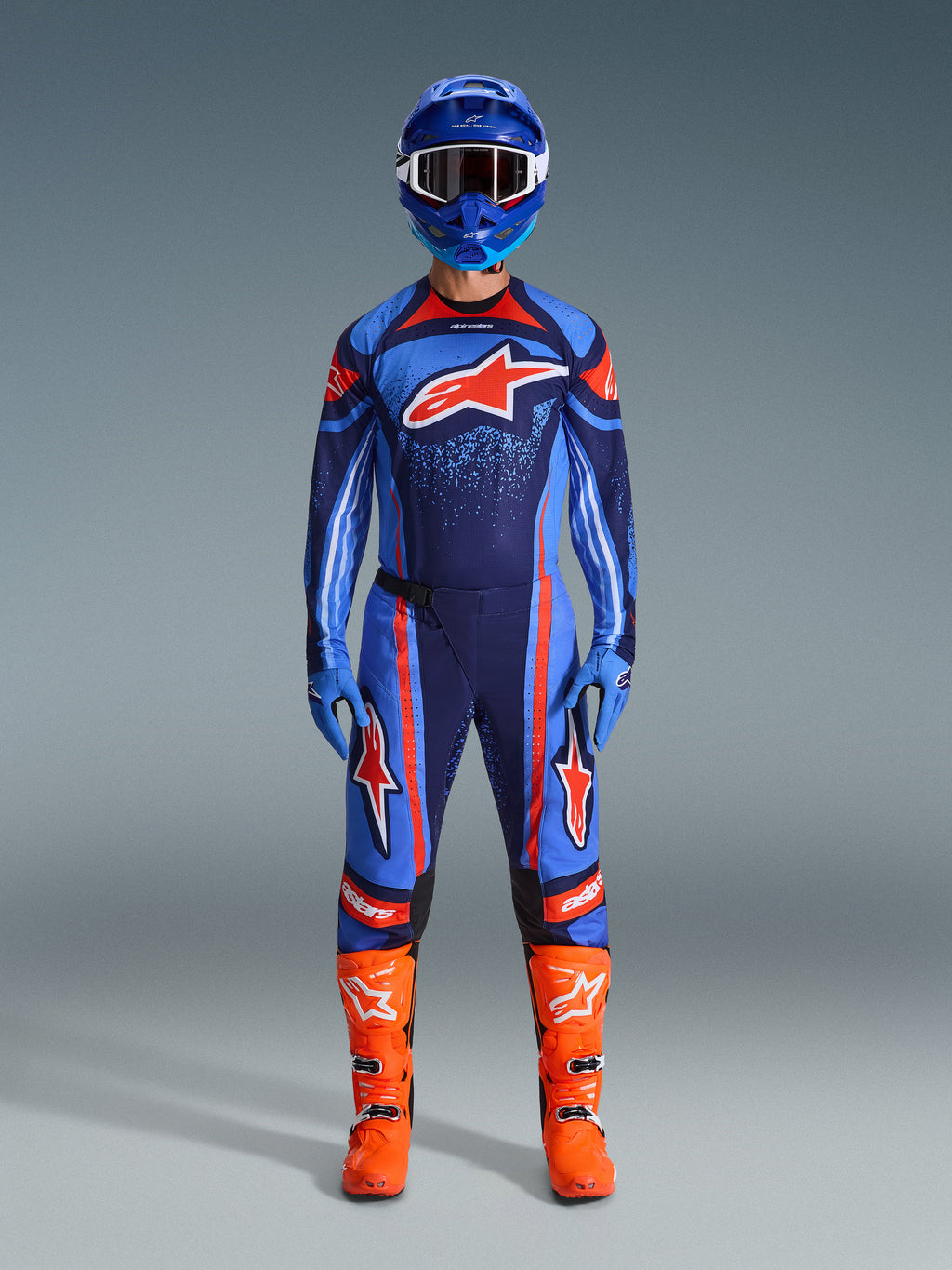 Alpinestars 2026 Techstar Nomur gear set, Motocross rider in full racing kit, Dark Navy with Orange and UCLA Blue accents, matching jersey and pants with large logos, blue helmet and bright orange boots, coordinated off-road competition gear.