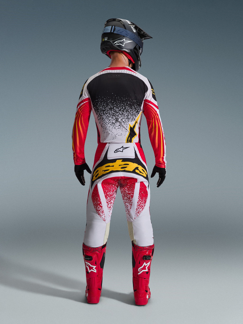 Rear view of a motocross rider wearing the Alpinestars 2026 Techstar Nomur Jersey in Black, Red, and White with yellow accents, paired with matching motocross pants featuring bold yellow branding, bright red racing boots, and a black helmet, complete coordinated gear set for off-road racing.