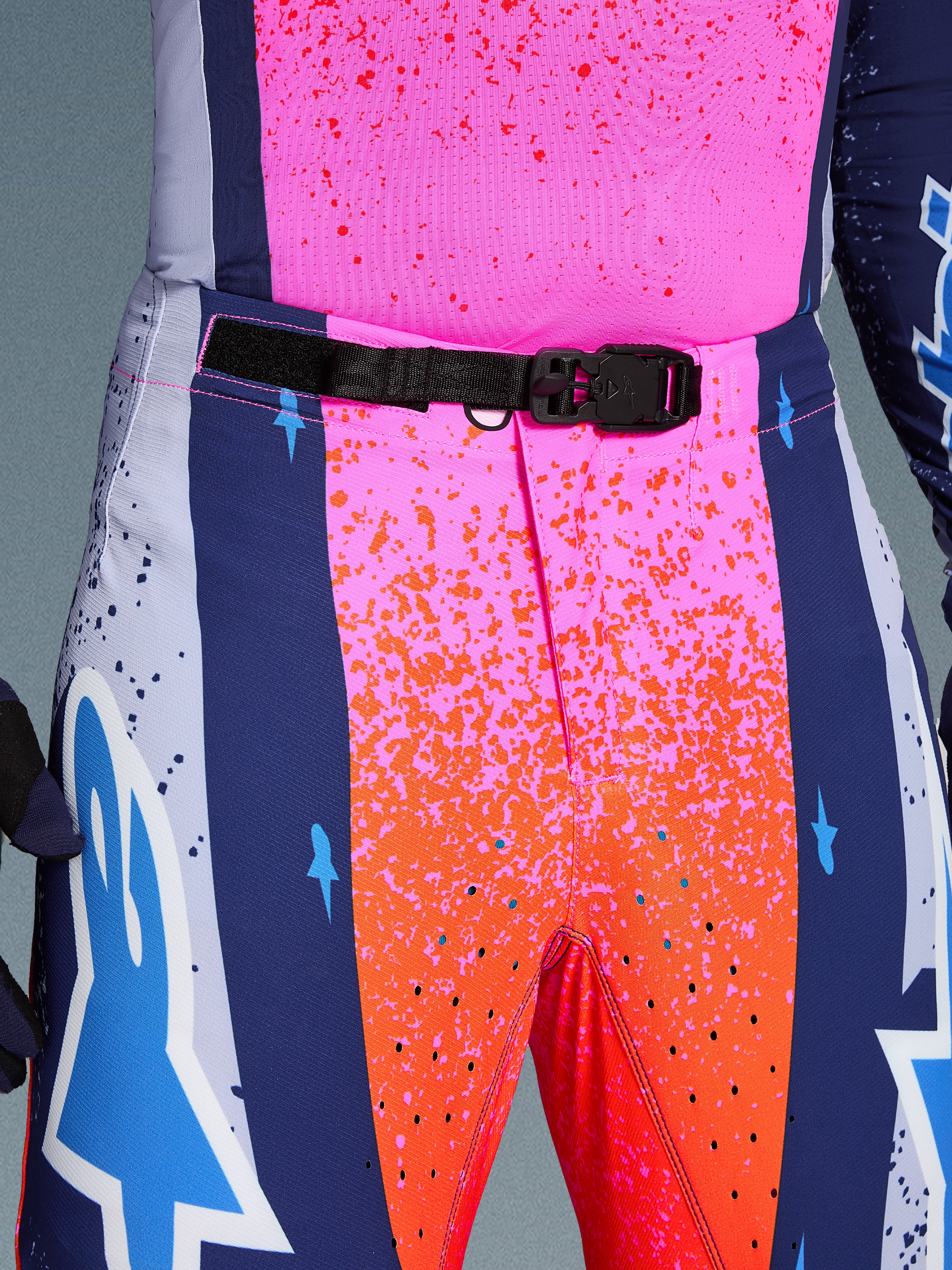 Alpinestars Limited Edition Supertech Records Pants, Motocross Pants, Multicolor pink and orange with navy blue side panels, close-up view of the waist featuring a black SX1 auto-locking magnetic buckle, rip-stop fabric with splatter graphics and laser-cut perforations.