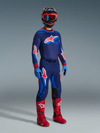 Motocross rider wearing 2026 Supertech Vista Pants in Dark Navy Hot Coral Cobalt, matching long-sleeve racing jersey, red motocross boots, and coordinated helmet with red goggles, full-body studio shot of high-performance off-road gear