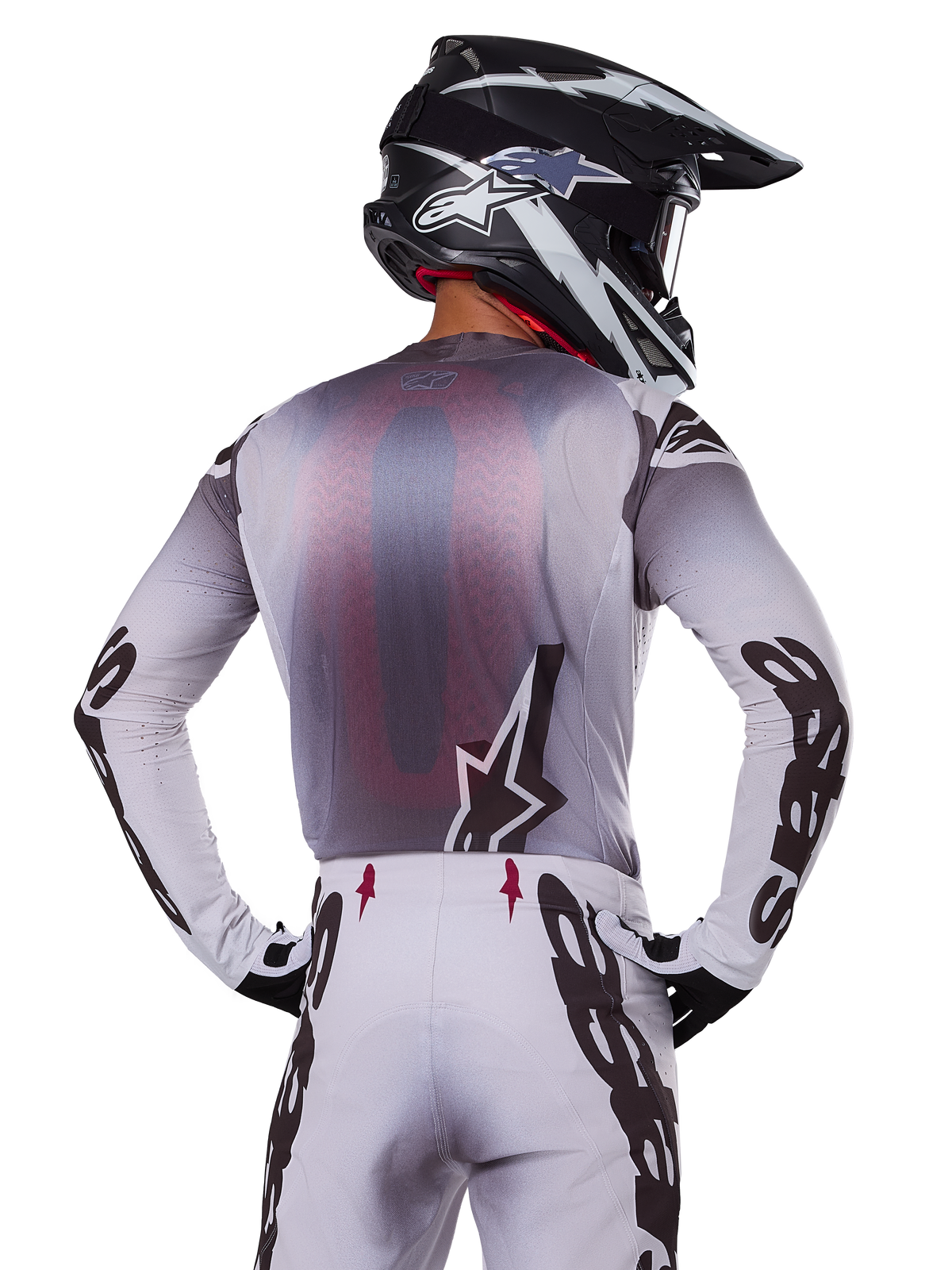 Motocross rider from behind wearing the Alpinestars 2025 Supertech Maker Jersey, Silver and Black with a red mesh back panel and black sleeve logos, paired with matching motocross pants and a black and white helmet, professional off-road racing gear