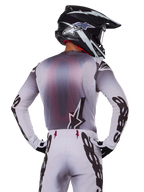 Motocross rider from behind wearing the Alpinestars 2025 Supertech Maker Jersey, Silver and Black with a red mesh back panel and black sleeve logos, paired with matching motocross pants and a black and white helmet, professional off-road racing gear