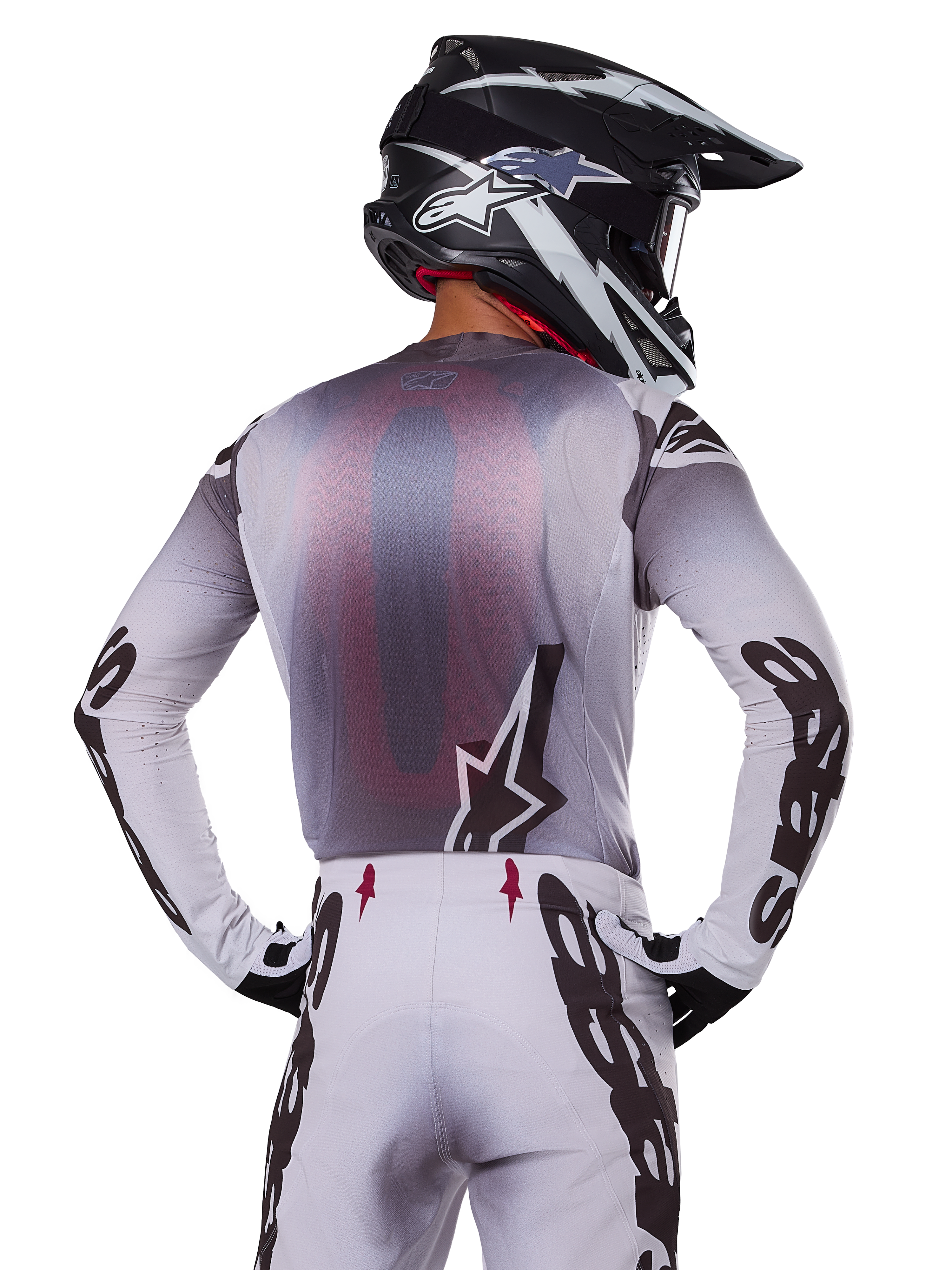 Motocross rider from behind wearing the Alpinestars 2025 Supertech Maker Jersey, Silver and Black with a red mesh back panel and black sleeve logos, paired with matching motocross pants and a black and white helmet, professional off-road racing gear