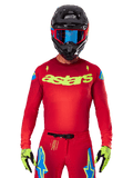 Alpinestars 2025 Supertech Maker Jersey in Bright Red Yellow Fluo, featuring yellow 'astars' chest branding and blue star shoulder designs, worn with matching pants, black helmet, blue goggles, and yellow gloves, professional motocross racing apparel