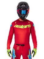 Alpinestars 2025 Supertech Maker Jersey in Bright Red Yellow Fluo, featuring yellow 'astars' chest branding and blue star shoulder designs, worn with matching pants, black helmet, blue goggles, and yellow gloves, professional motocross racing apparel