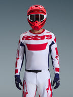 Motocross rider wearing the Alpinestars 2026 Racer Riway Jersey in White Blue Red, featuring a large red 'astars' logo on the chest, navy blue side panels, matching white and red motocross pants, a red helmet with white goggles, and navy blue gloves, complete off-road racing gear set.