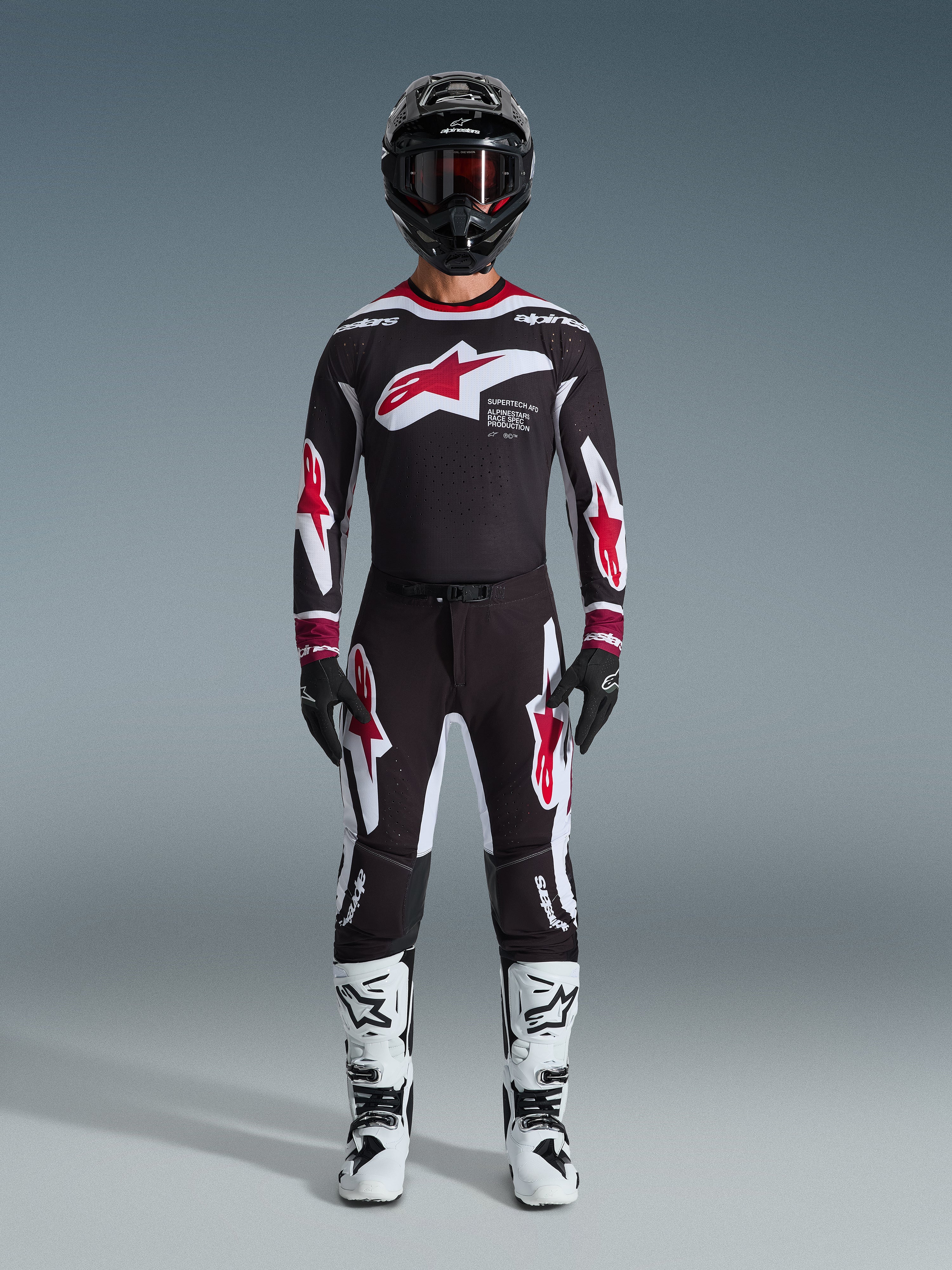 Motocross rider wearing Alpinestars 2026 Supertech AFD Vista Jersey, Black Red and White racing gear, matching black pants with red star logos, white and black boots, black helmet with goggles, professional off-road racing kit.