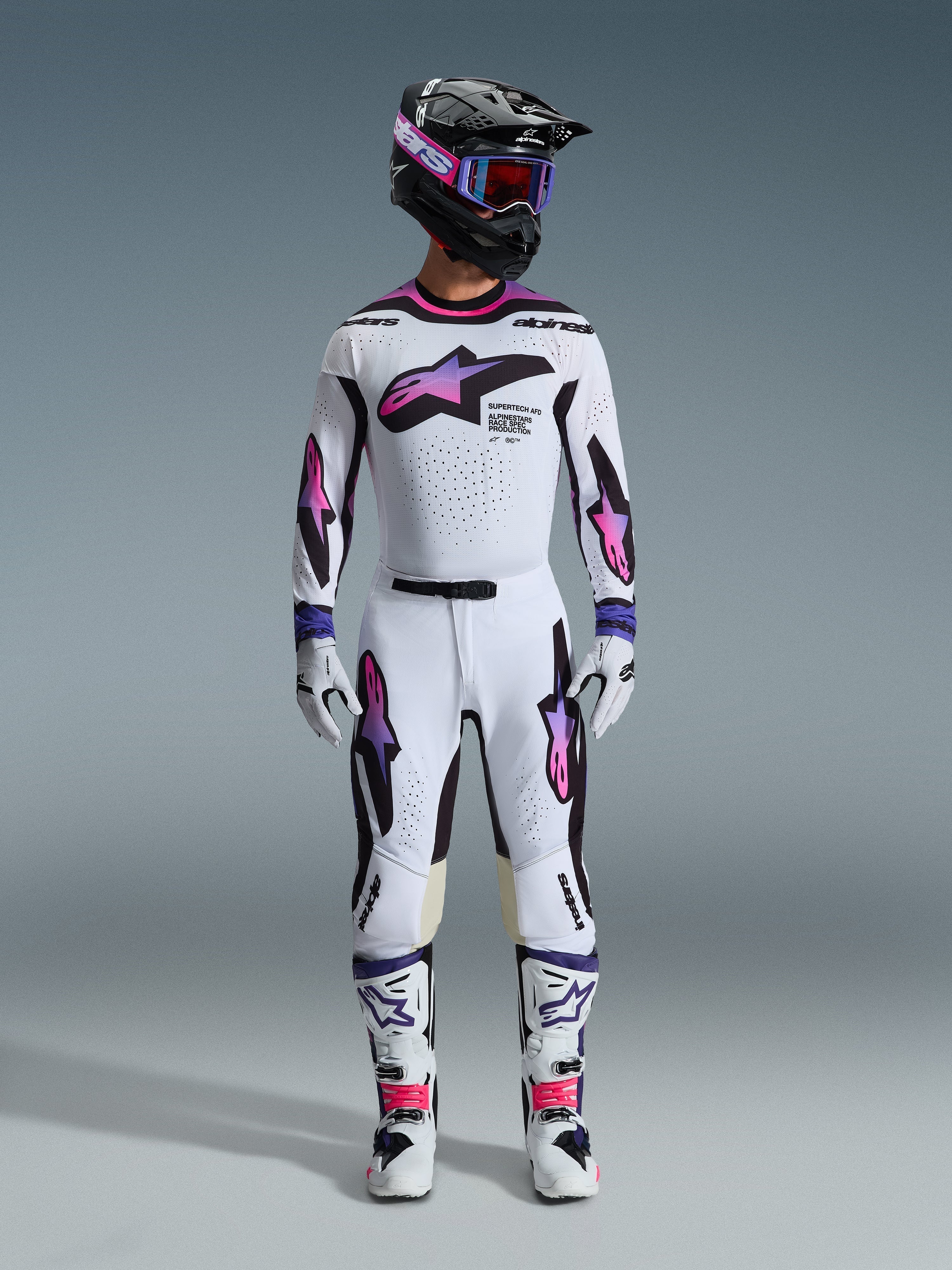 Motocross rider wearing complete racing gear, Alpinestars 2026 Supertech AFD Vista Jersey in White Purple Black, matching white pants with purple and pink gradient graphics, white boots, and black helmet with purple goggles, professional off-road racing outfit