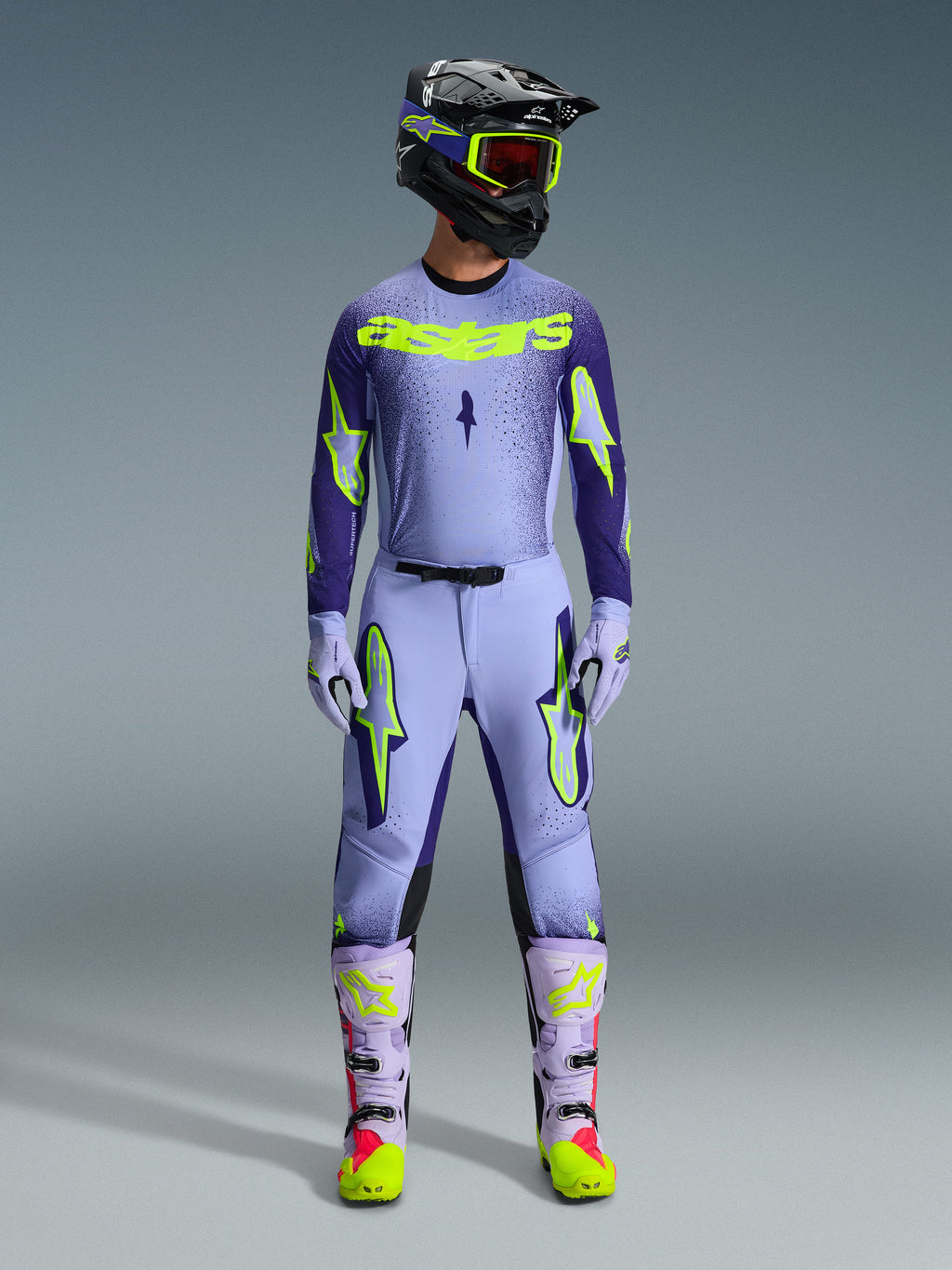 Motocross rider wearing complete Alpinestars 2026 Supertech Scenz racing gear, Purple and Yellow Fluo jersey with matching pants, black helmet with yellow goggles, light purple and neon boots, professional athletic-fit outfit for off-road racing