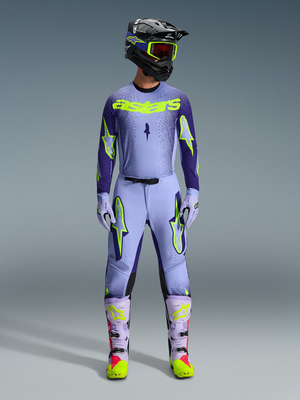 Motocross rider wearing complete Alpinestars 2026 Supertech Scenz racing gear, Purple and Yellow Fluo jersey with matching pants, black helmet with yellow goggles, light purple and neon boots, professional athletic-fit outfit for off-road racing