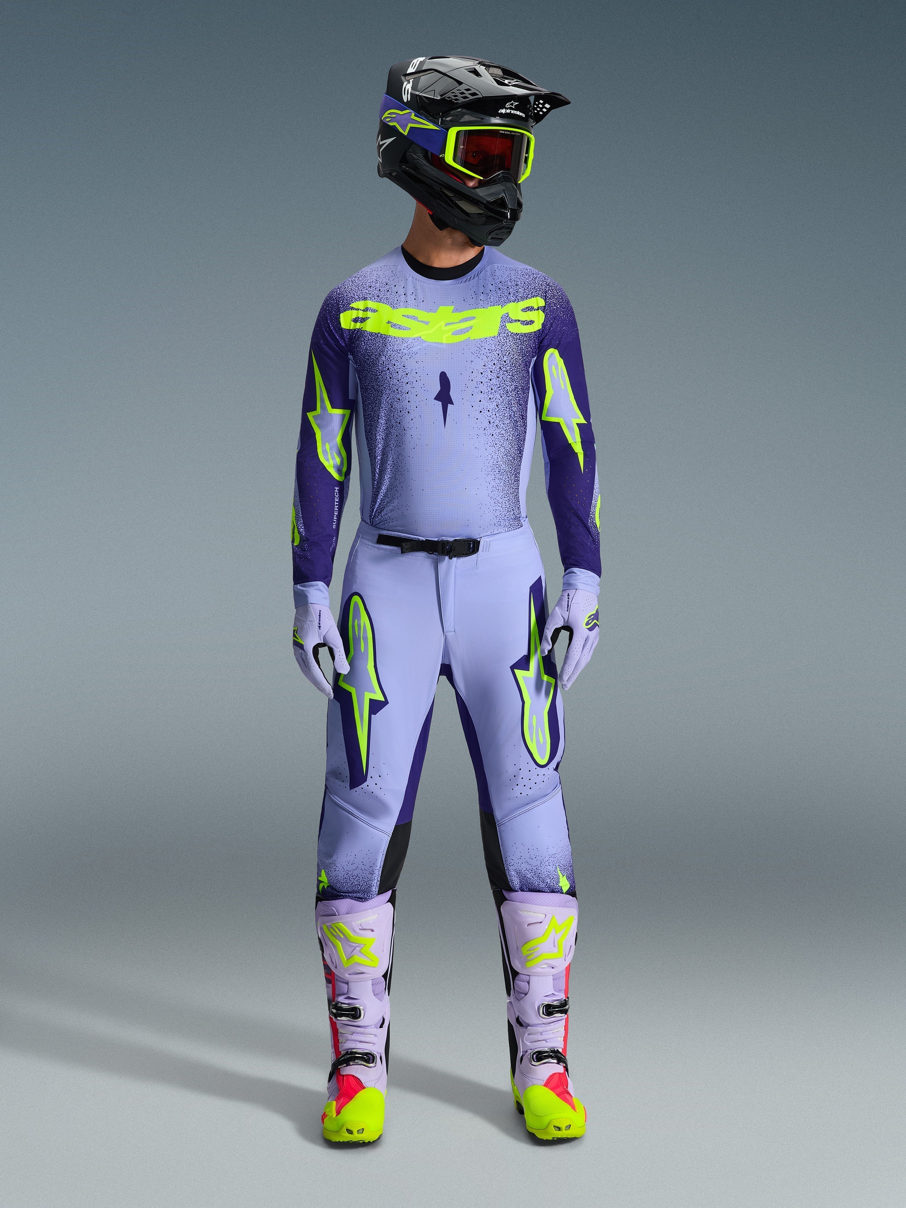 Motocross rider wearing complete Alpinestars 2026 Supertech Scenz racing gear, Purple and Yellow Fluo jersey with matching pants, black helmet with yellow goggles, light purple and neon boots, professional athletic-fit outfit for off-road racing