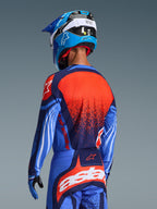Motocross rider from behind wearing the 2026 Techstar Nomur Jersey, Dark Navy with Orange and UCLA Blue accents, featuring a large orange textured graphic on the back, paired with matching motocross pants and a blue racing helmet, coordinated professional gear for off-road riding.