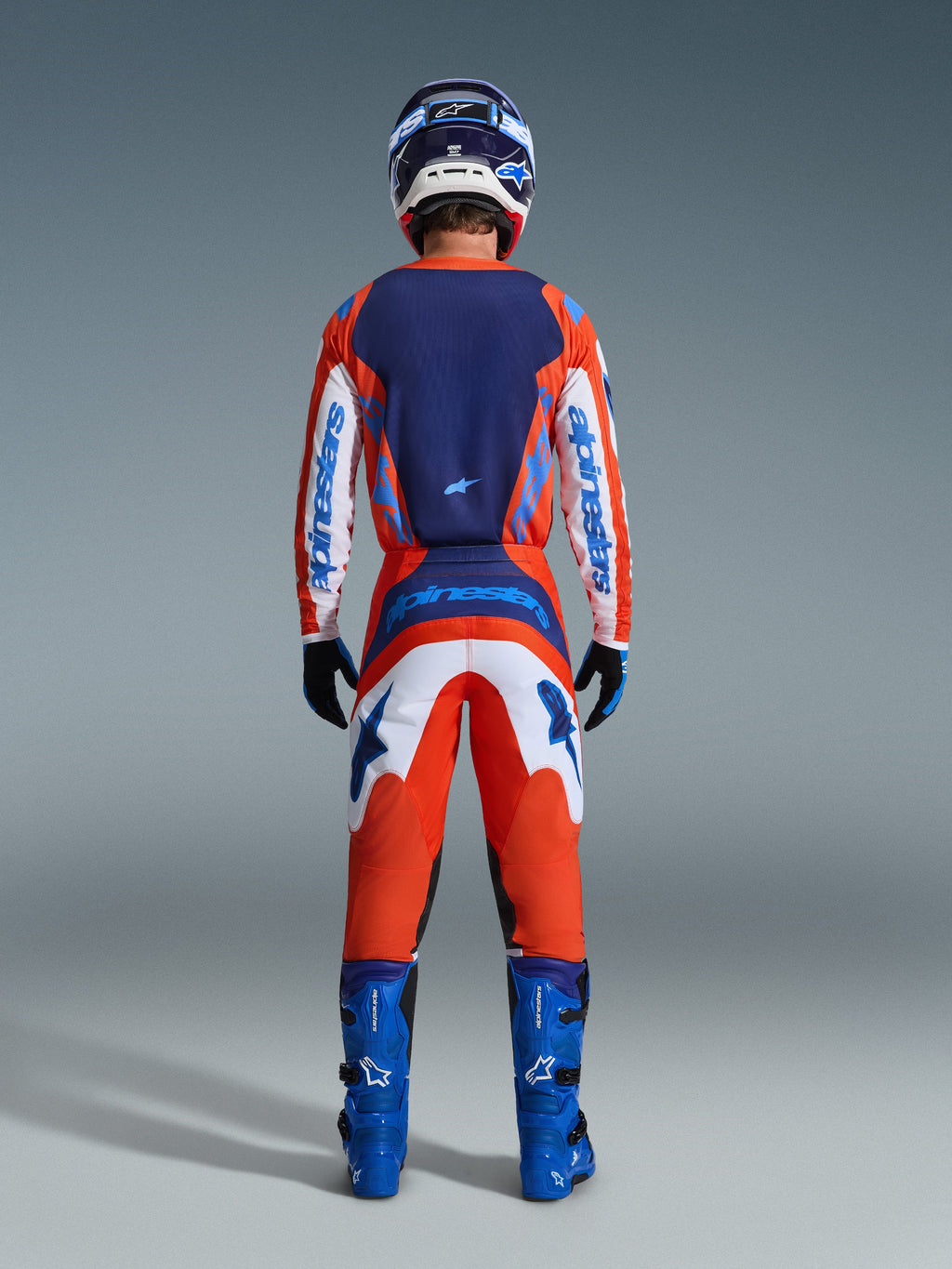 Alpinestars 2026 Fluid Grid Jersey, Motocross Jersey, Orange and UCLA Blue, back view of rider in full racing gear set, featuring a dark blue back panel and white sleeves with blue branding, paired with matching pants and blue boots.