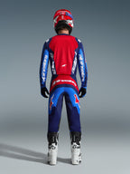 Rear view of a motocross rider wearing the 2026 Fluid Grid Jersey in Blue and Red, featuring a large red back panel and blue sleeves with white Alpinestars branding, paired with matching pants, white and black boots, and a red helmet for a complete off-road racing outfit.
