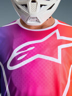 Alpinestars 2026 Fluid Apex Jersey, Motocross Jersey, Multicolor Purple Pink and Orange Gradient, featuring a large white Astars logo on the chest, close-up view worn with a white motocross helmet