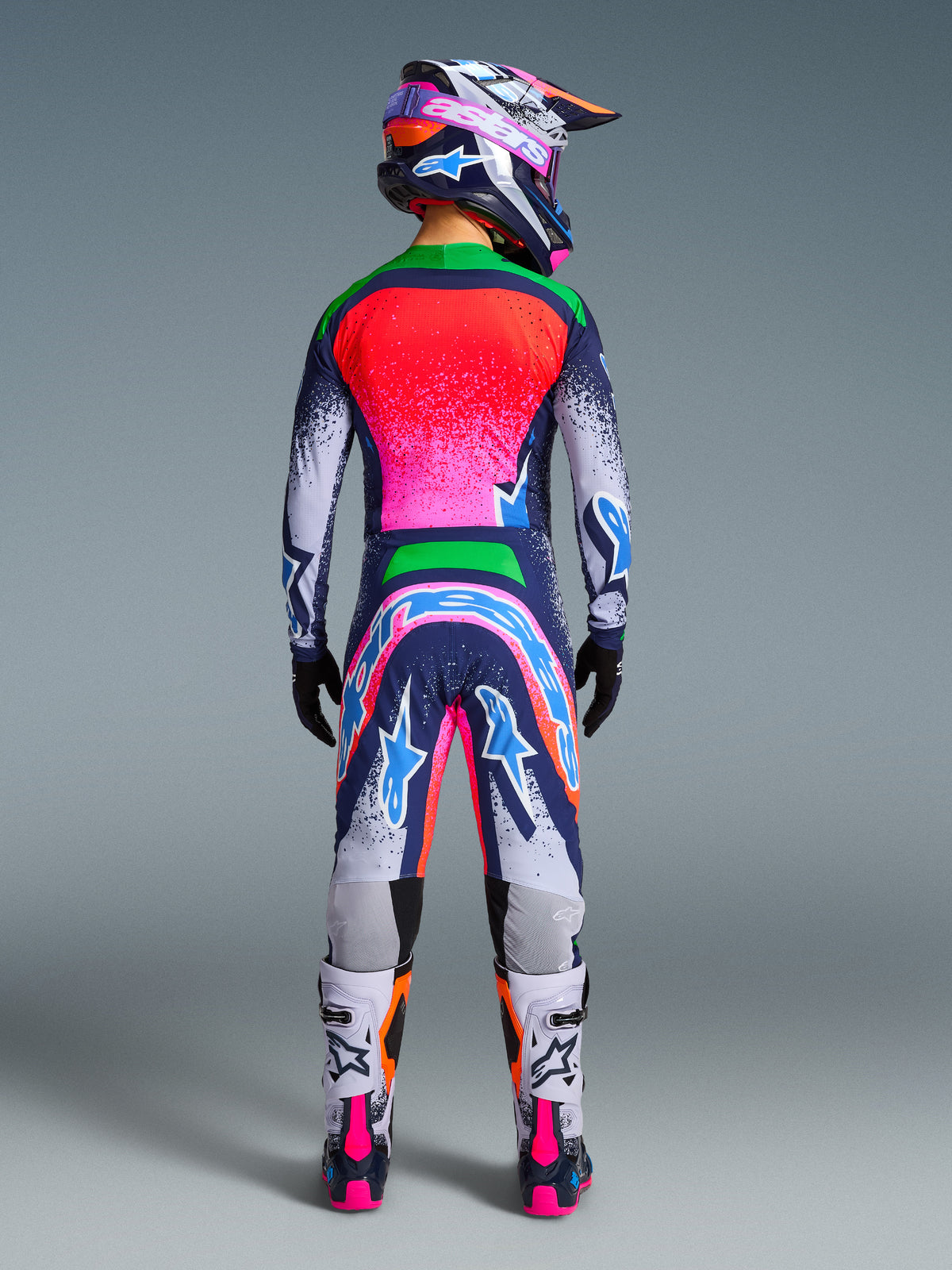Motocross rider seen from the back wearing the Alpinestars Limited Edition Supertech Records Gear Set, vibrant multicolor jersey and pants with pink, orange, and blue splatter design, matching helmet, black gloves, and white and pink boots, professional off-road racing gear.