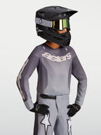 Alpinestars 2027 Youth Fluid Amber Jersey, youth motocross jersey in sand and gray, on-model with a black racing helmet.