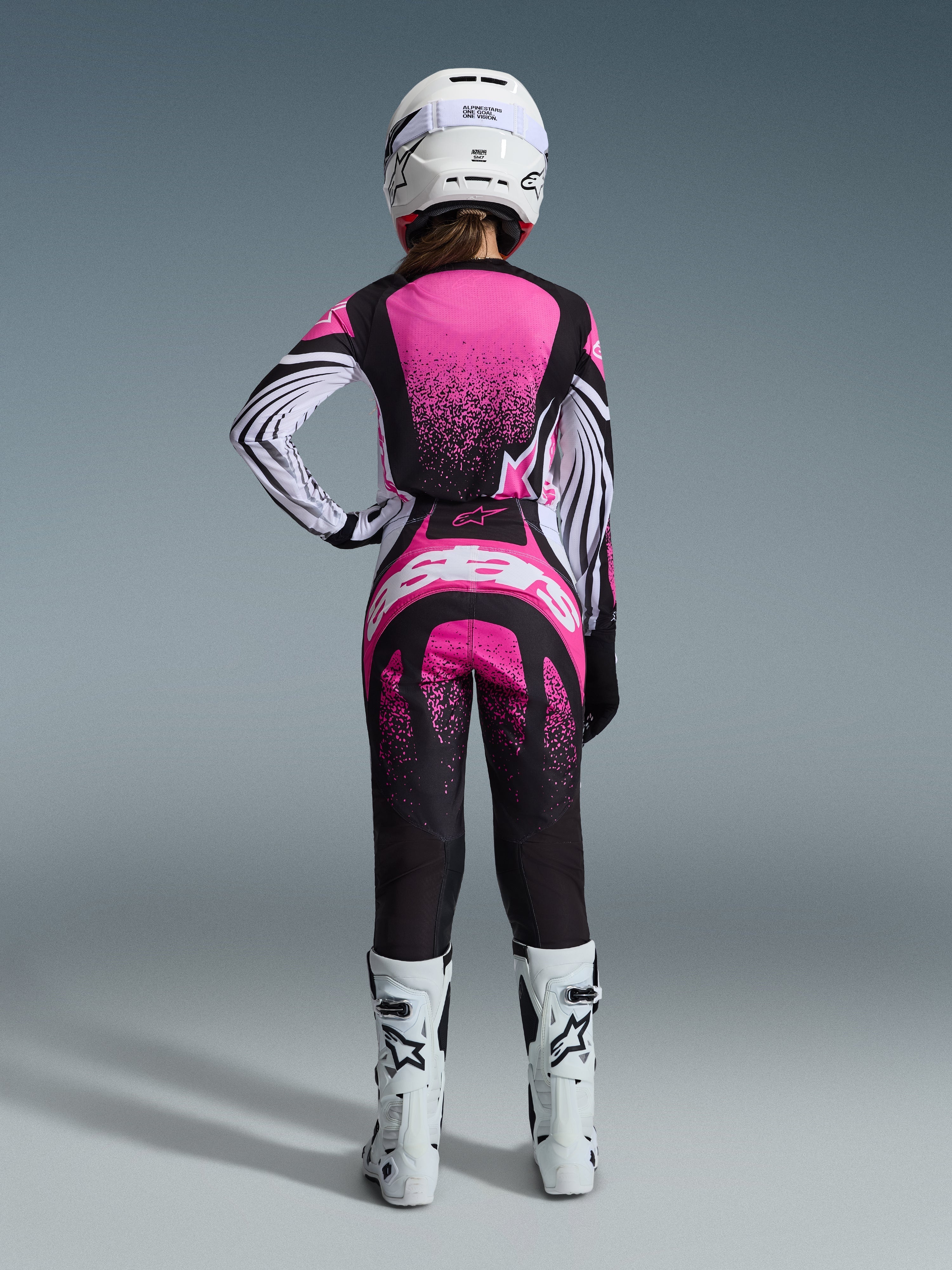 Alpinestars 2026 Stella Techstar Nomur Gear Set, female motocross rider viewed from behind, pink and black jersey with white striped sleeves and speckled pattern, matching pants with white Alpinestars logo, white helmet and white off-road boots