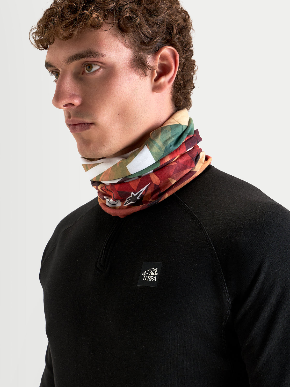 Alpinestars Buff Peaks Primaloft Neck Tube, earth brown and multi-color printed neckwear worn by a model, featuring low-bulk PrimaLoft insulation and seamless tubular knit construction for all-terrain motorcycle riding