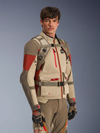 Alpinestars AT-15 Back Pack in Aluminum Walnut, worn by a rider over a matching adventure motorcycle jacket, showing the front harness with magnetic chest lock and adjustable straps, tan and red technical gear for adventure riding