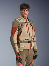 Alpinestars AT-15 Back Pack in Aluminum Walnut, worn by a rider over a matching adventure motorcycle jacket, showing the front harness with magnetic chest lock and adjustable straps, tan and red technical gear for adventure riding