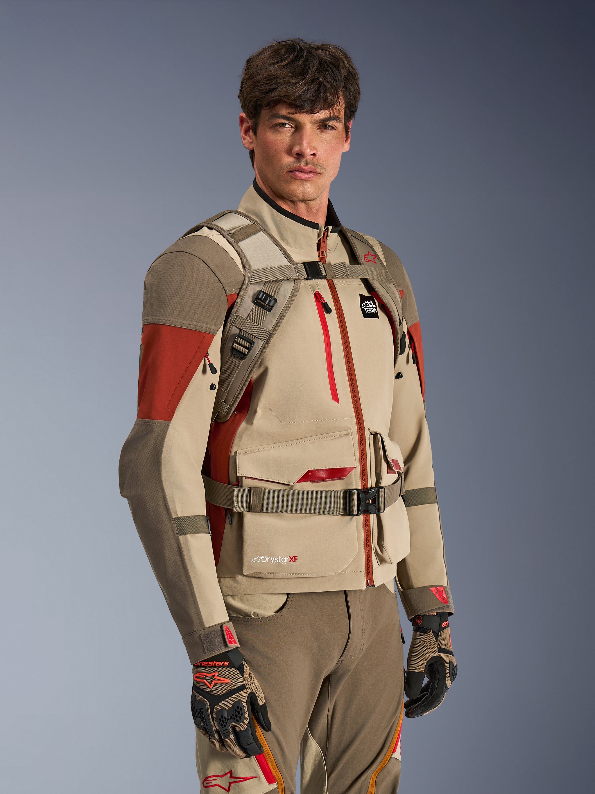 Alpinestars AT-15 Back Pack in Aluminum Walnut, worn by a rider over a matching adventure motorcycle jacket, showing the front harness with magnetic chest lock and adjustable straps, tan and red technical gear for adventure riding