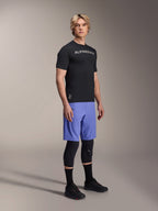Male model wearing Alpinestars A-Aria Switch Short Sleeve Jersey in Black with white chest branding, paired with light blue mountain bike shorts, black knee guards, and black cycling shoes, studio full-body shot for performance MTB gear