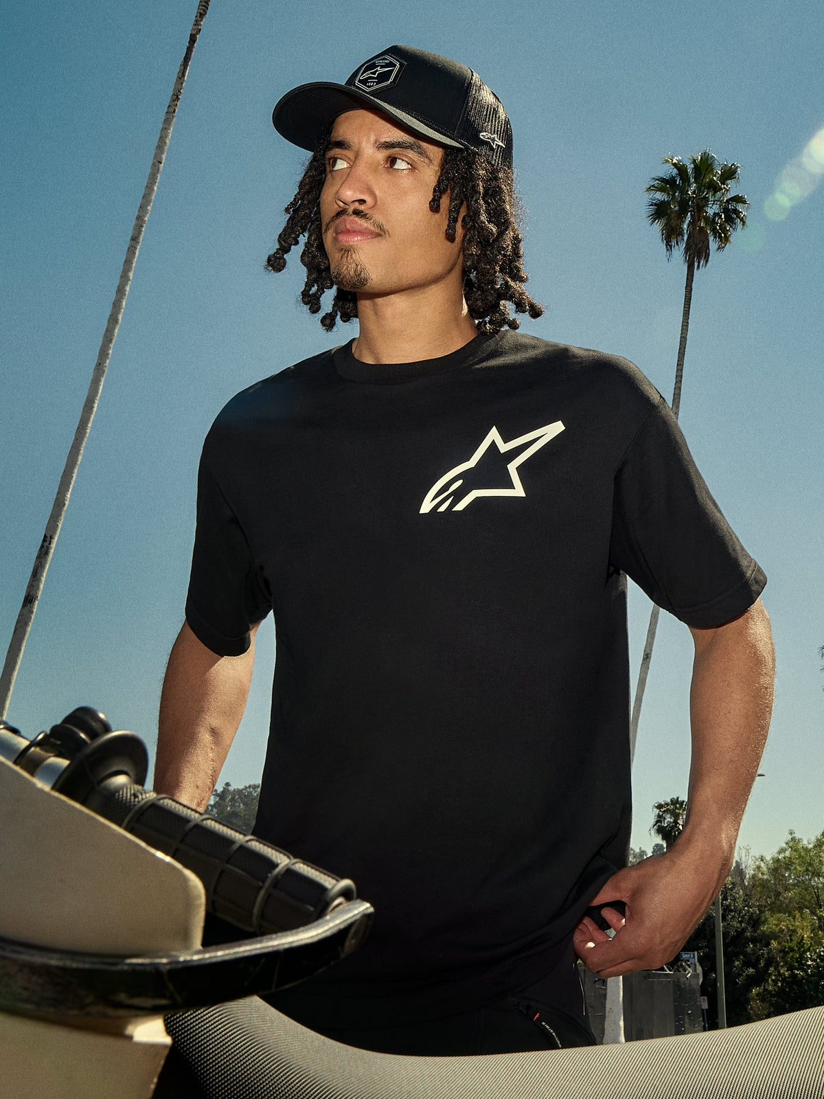 Alpinestars One Oversized CSF Tee, Short Sleeve T-Shirt, Black with white Astars logo, worn by a man in a black trucker hat, outdoor lifestyle setting with palm trees and a motorcycle, casual motorsport streetwear