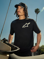 Alpinestars One Oversized CSF Tee, Short Sleeve T-Shirt, Black with white Astars logo, worn by a man in a black trucker hat, outdoor lifestyle setting with palm trees and a motorcycle, casual motorsport streetwear