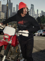 Alpinestars Pulse Hoodie, Black, hooded pullover fleece with red and grey Astars Combo Logo, worn by a man in an orange beanie standing next to a red Honda motorcycle in a city setting, casual motorsport streetwear