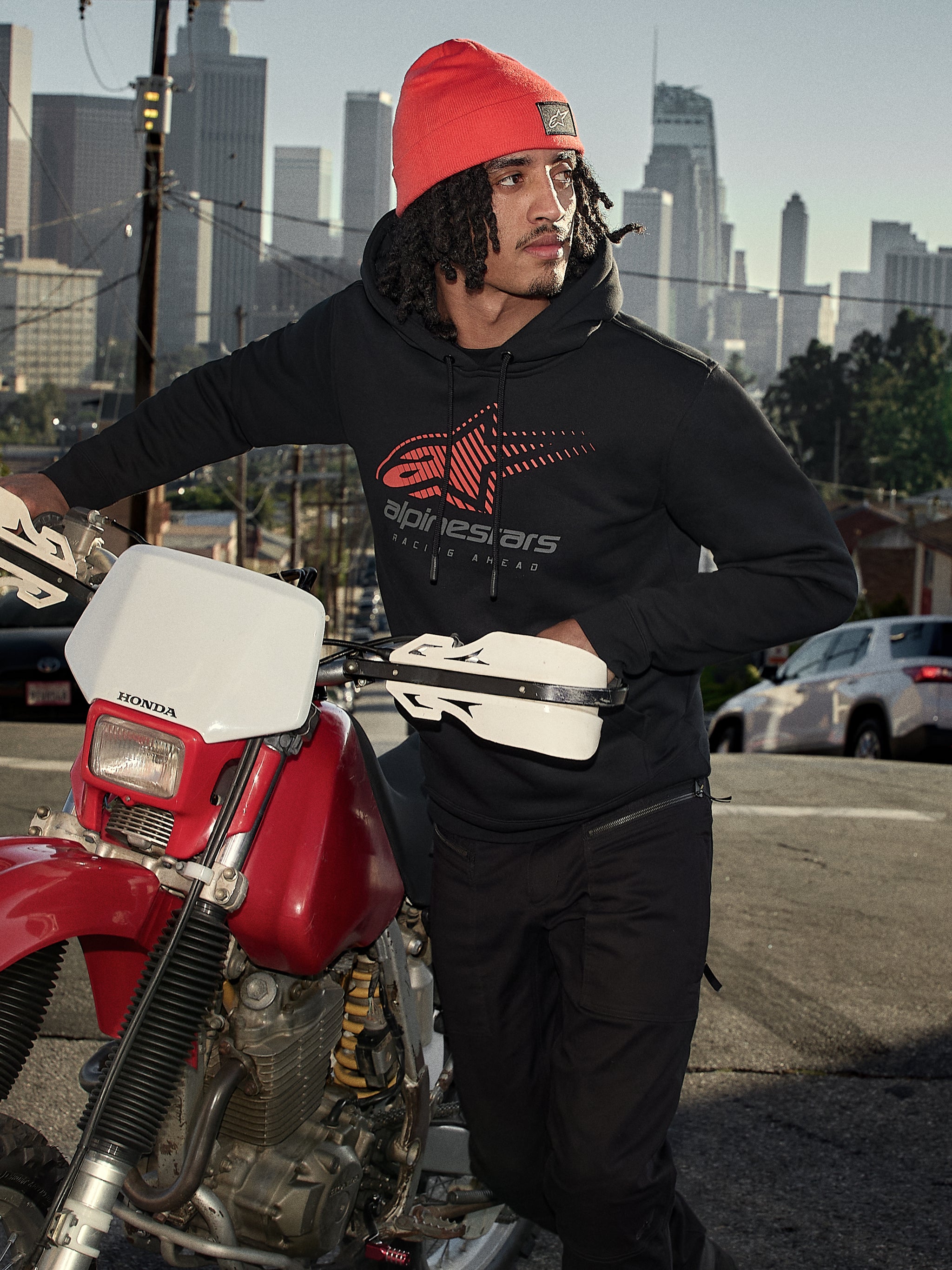 Alpinestars Pulse Hoodie, Black, hooded pullover fleece with red and grey Astars Combo Logo, worn by a man in an orange beanie standing next to a red Honda motorcycle in a city setting, casual motorsport streetwear