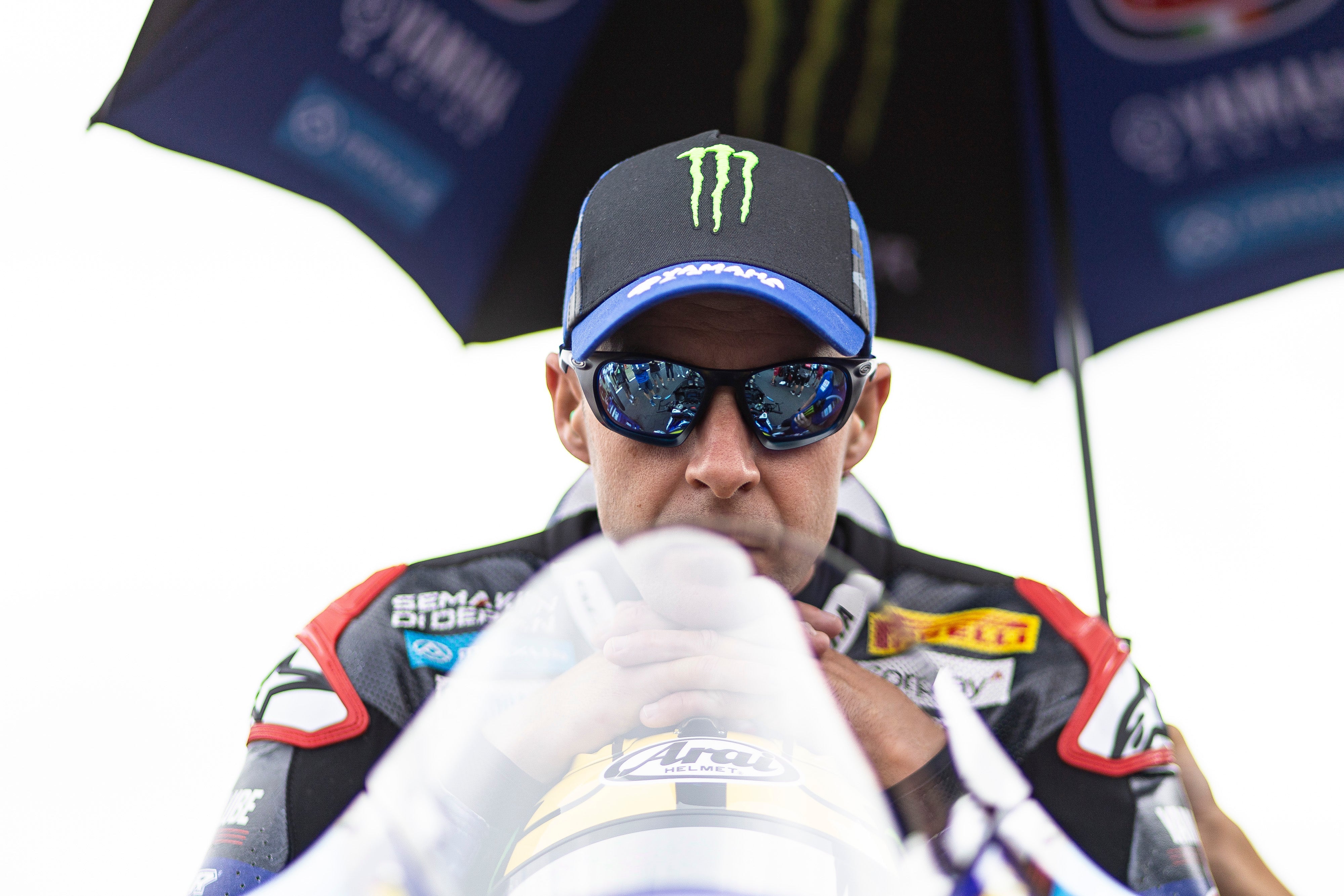 Jonathan Rea wearing Alpinestars racing suit and sunglasses under an umbrella with visible branding.